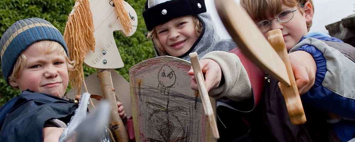 Drei als Ritter verkleidete Kinder, die Holzschwerter und Schilde halten. Auf einem der Schilde ist eine Zeichnung eines Skeletts zu sehen.