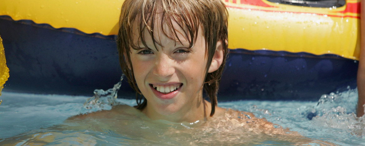 Ein Kind in einem Schwimmbecken, wobei ihm das Wasser bis zu den Schultern reicht. Im Hintergrund ist eine gelb-blaue Luftmatratze zu sehen.