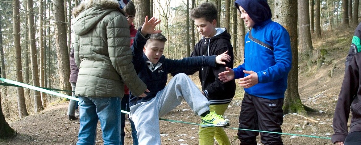 Eine Gruppe von Kindern im Wald, wobei ein Kind versucht auf eine Slackline aufzusteigen und zu balancieren, während die anderen in der Nähe stehen und ihm dabei helfen.