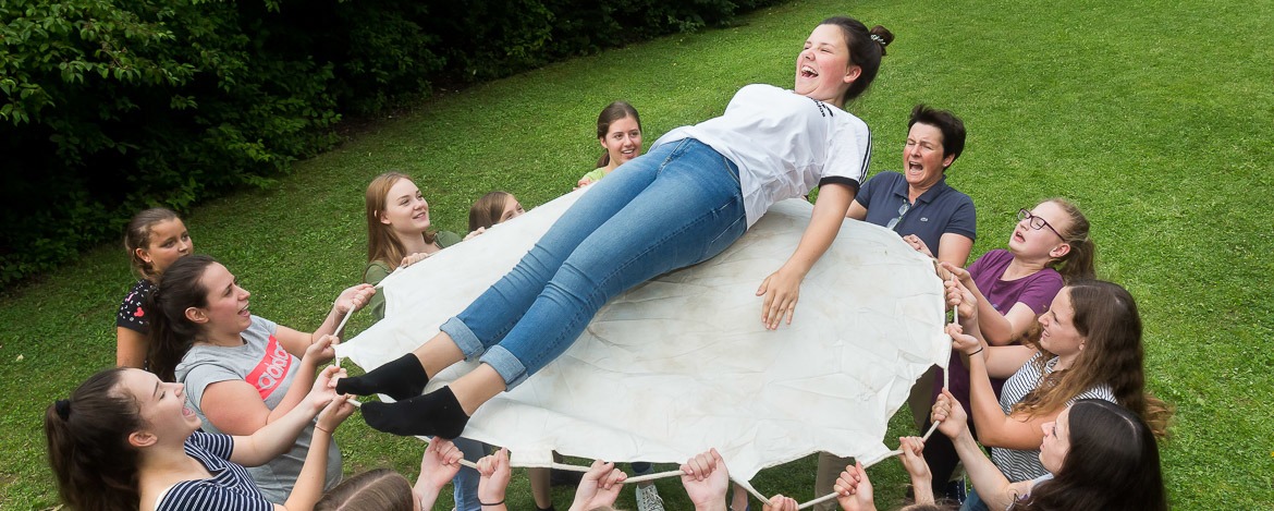 Eine Gruppe von Mädchen, die ein Mädchen auf einem großen runden Stück Stoff halten. Das Mädchen wird von der Gruppe in einer Außenumgebung mit Gras und Bäumen im Hintergrund hochgehoben.