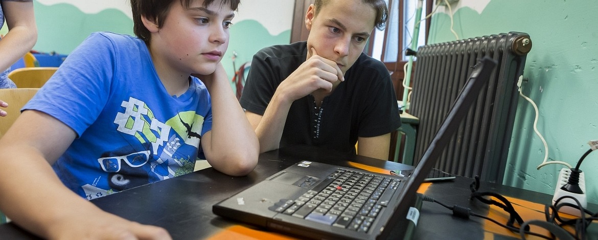 Zwei Jungs, die an einem Tisch sitzen, auf dem sich ein Laptop befindet. Sie konzentrieren sich auf den Inhalt des Laptop-Bildschirms.
