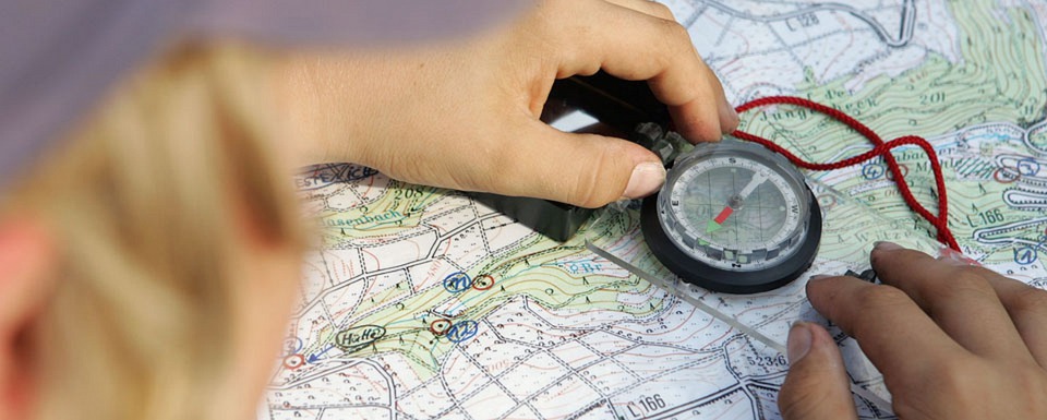 Eine Person, die eine Karte anschaut und einen Kompass benutzt. Die Hände der Person sind auf der Karte platziert, wobei der Kompass auf einem Teil der Karte liegt.