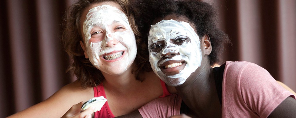 Zwei junge Frauen, die eine weiße Gesichtsmaske im Gesicht haben. Die Person links hat die Gesichtscreme ebenfalls in der Hand.