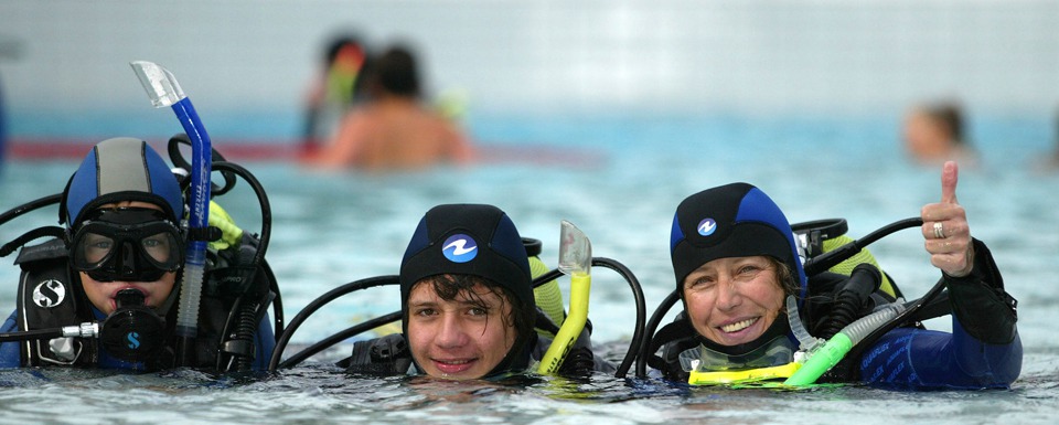 Drei Personen in Tauchausrüstung, die teilweise im Wasser sind, wobei eine Person einen Daumen-hoch-Gestus macht. Im Hintergrund sind weitere Personen zu sehen, die ebenfalls im Wasser sind.