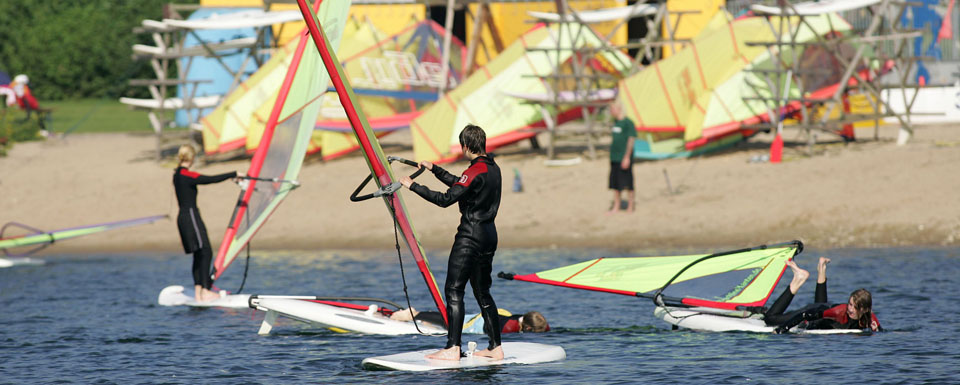 Mehrere Personen in Neoprenanzügen, die auf Windsurfbrettern auf einem Gewässer stehen. Im Hintergrund sind bunte Segel und eine sandige Küste zu sehen.